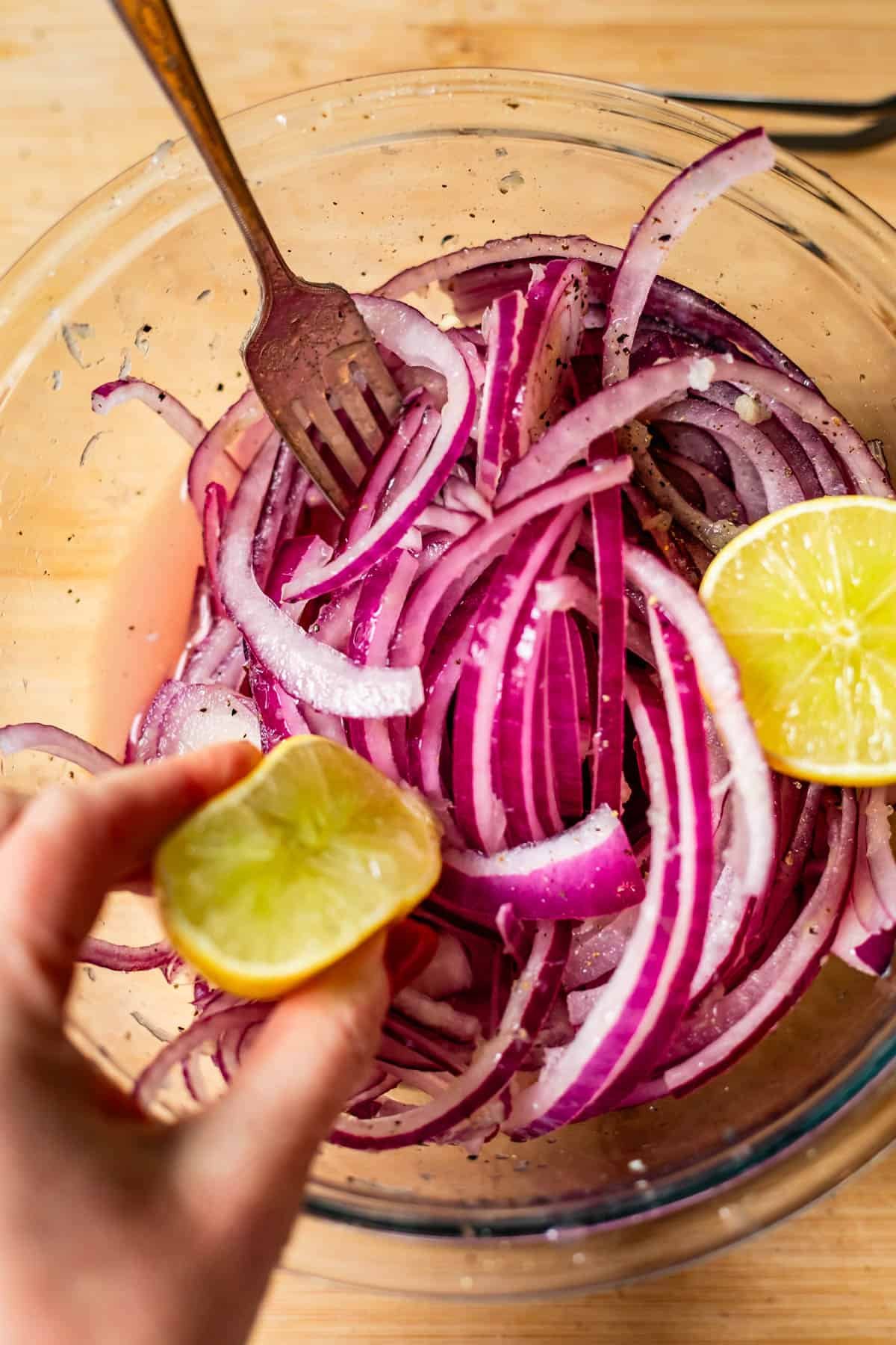 A hand squeezes a halved lime over a bowl of sliced red onions, with a fork in the bowl, on a wooden surface. The onions are seasoned with pepper and other visible spices.