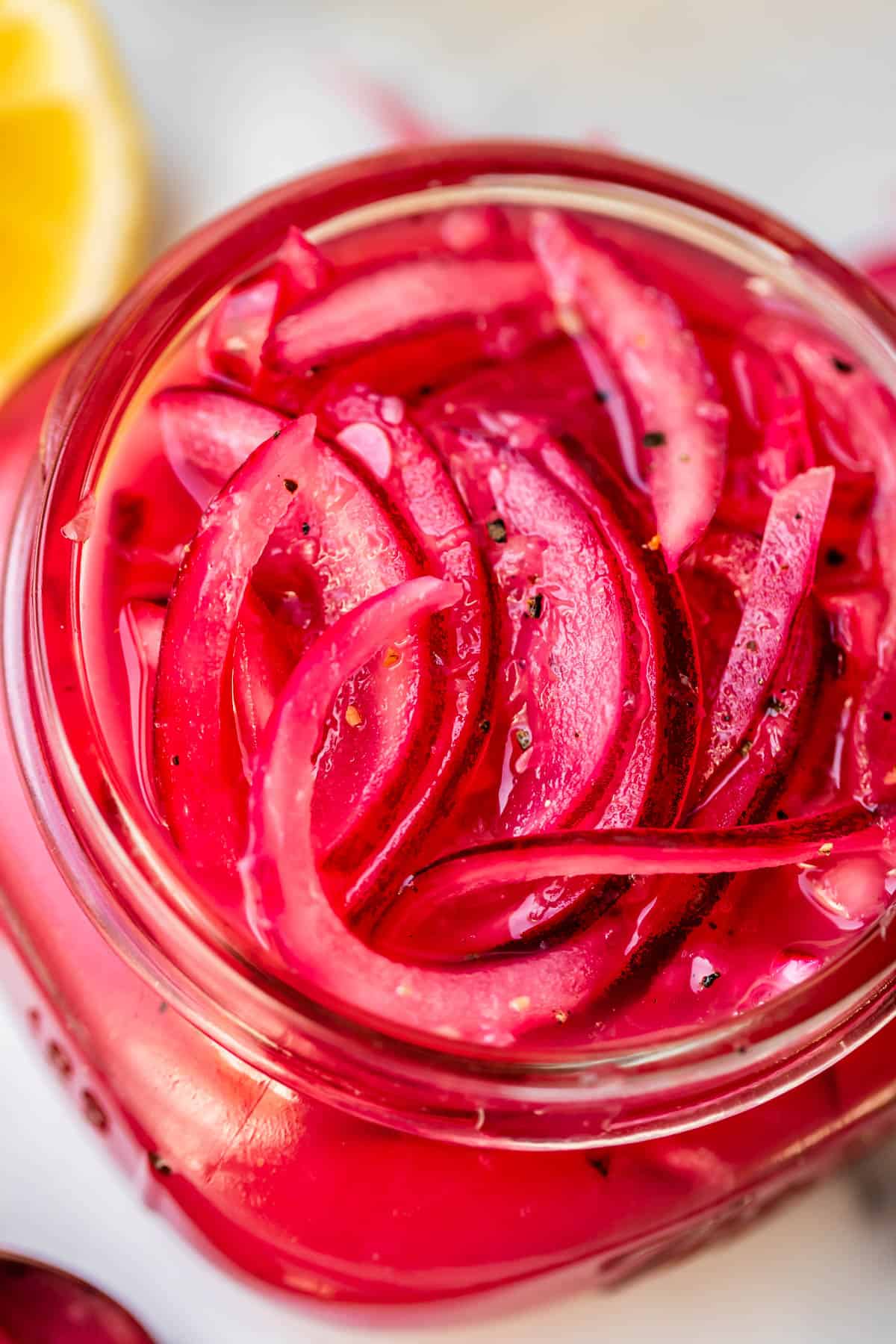 Close-up of vibrant pink pickled red onion slices in a glass jar, with a partial lemon wedge visible in the background. The onions are thinly sliced and soaked in brine.