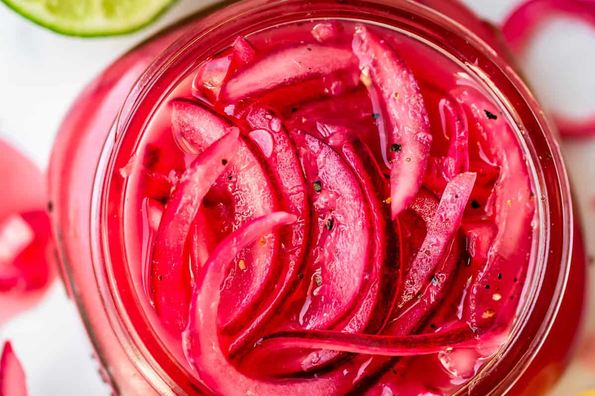 Close-up top view of thinly sliced pickled red onions in a glass jar, with visible black pepper specks and a partial lime slice in the corner. The onions are bright pink and glossy.