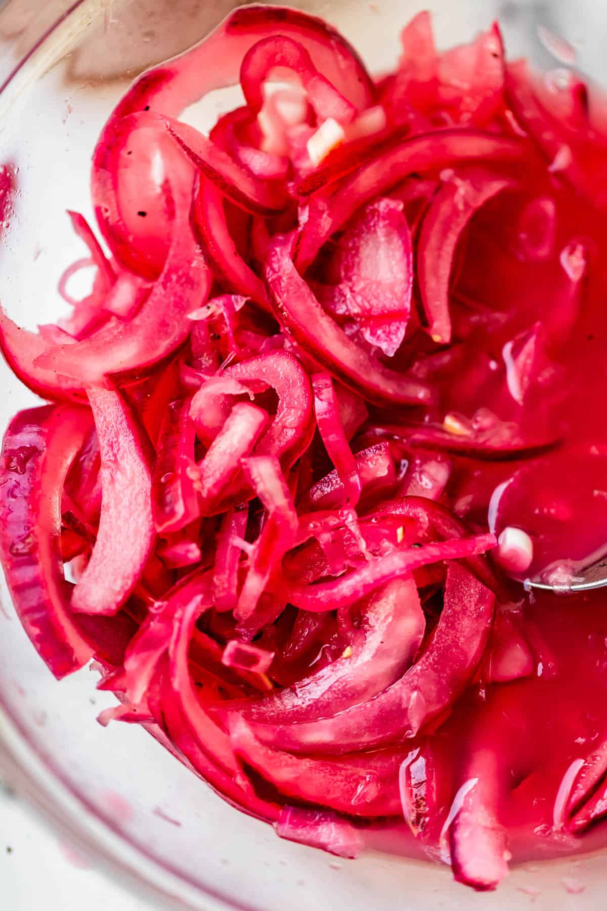 Thinly sliced red onions in a pinkish pickling liquid, shown in close-up inside a clear glass bowl. The onions have a vibrant magenta color and appear slightly translucent.