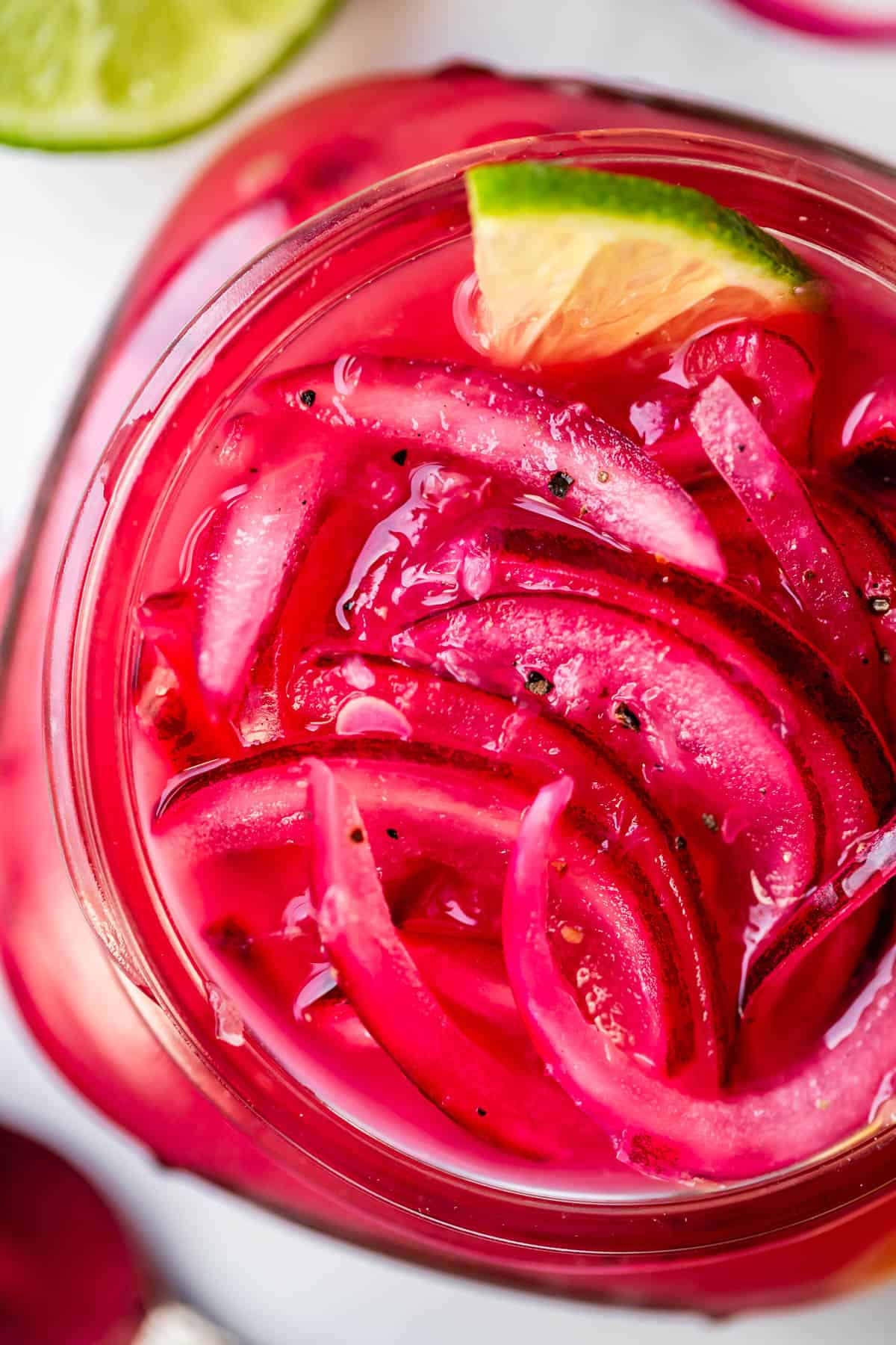 Close-up of pickled red onions in a jar, garnished with a wedge of lime. The onions are submerged in a bright pink brine, and the image is vibrant with strong colors and glossy textures.