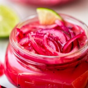 A glass jar filled with vibrant pink pickled red onions, garnished with a lime wedge. Slices of lime and a spoon are nearby, and another jar of pickled onions is blurred in the background.