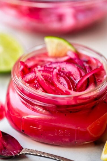A glass jar filled with vibrant pink pickled red onions, garnished with a lime wedge. Slices of lime and a spoon are nearby, and another jar of pickled onions is blurred in the background.