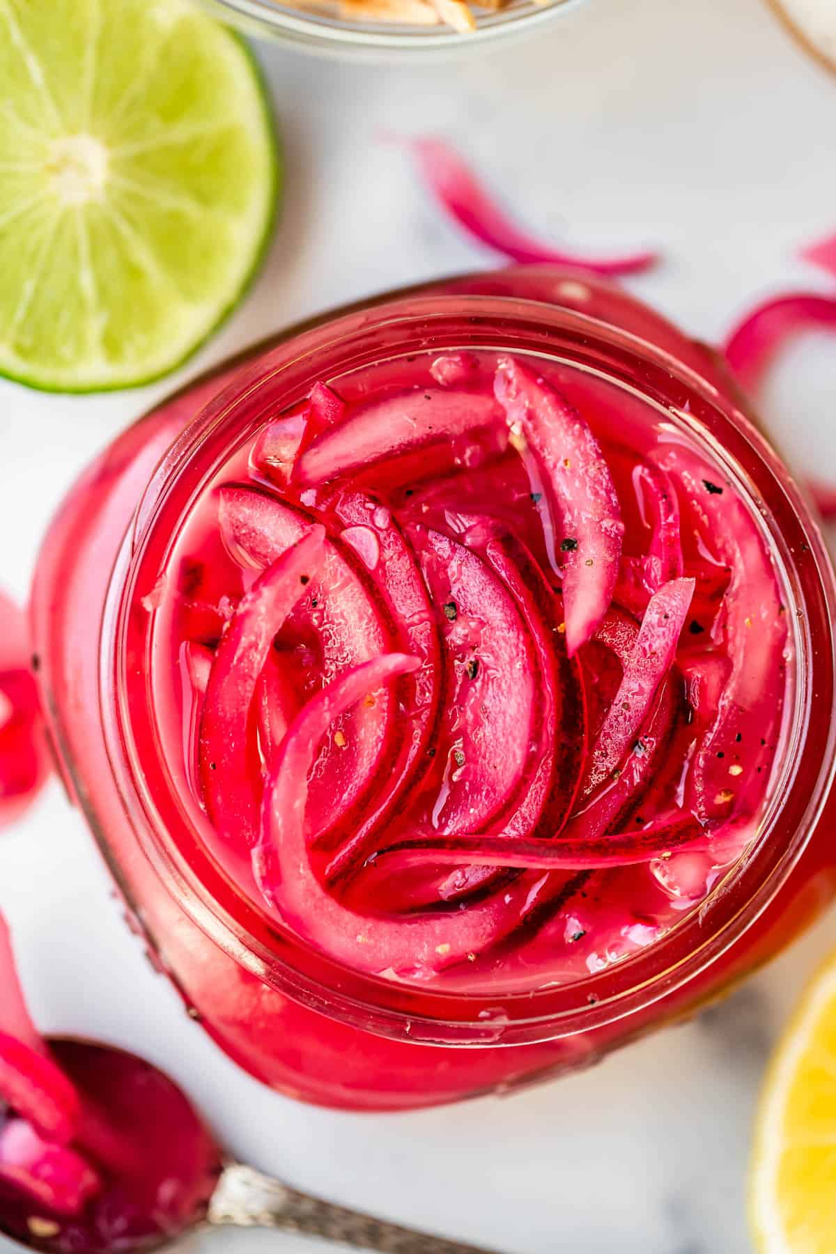 A close-up top view of a glass jar filled with vibrant pink pickled red onions, sprinkled with black pepper. Slices of lime are visible nearby on a white surface.