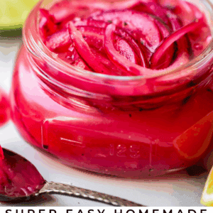 A small glass jar filled with bright pink pickled red onions, garnished with a lime wedge, sits on a white surface. Text below reads: Super Easy Homemade Pickled Red Onions - The Food Charlatan.