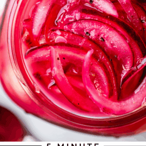 A close-up of a jar filled with bright pink pickled red onions, garnished with a lime wedge. Text below reads 5 Minute Pickled Red Onions - The Food Charlatan.