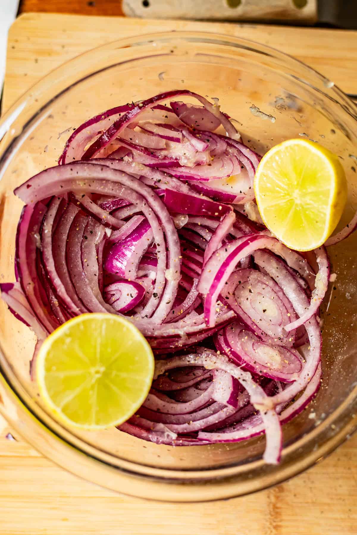 A glass bowl with thinly sliced red onions, black pepper, and salt, garnished with two halves of a lemon, placed on a wooden surface.
