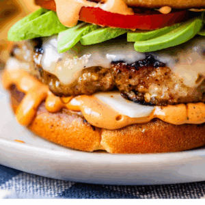Close-up of a turkey burger with melted cheese, sliced avocado, tomato, and creamy sauce on a toasted bun. Text at the bottom reads, “Turkey Burgers That Don’t Suck – The Food Charlatan.”.