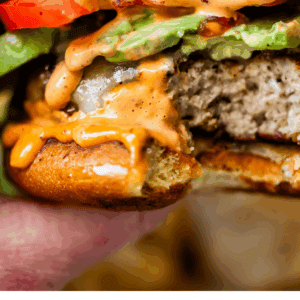 A close-up of a hand holding a turkey burger with lettuce, tomato, and a creamy orange sauce, showing a bite taken out. Text below reads, “Simple Homemade Turkey Burgers - The Food Charlatan.”.