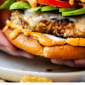 A close-up of a turkey burger with melted cheese, avocado slices, tomato, and sauce in a bun, held in a hand. Text below reads: Stupid Easy Turkey Burgers - The Food Charlatan.