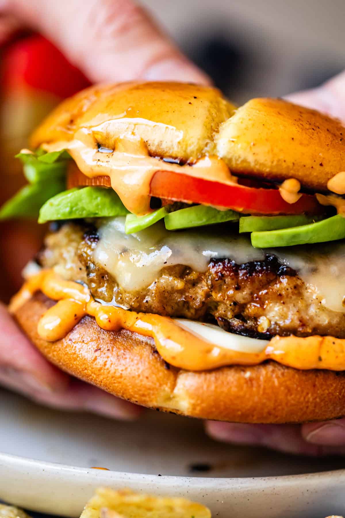 A close-up of hands holding a cheeseburger with melted cheese, lettuce, tomato, creamy sauce, and a glossy toasted bun. The burger is juicy with sauce dripping onto the plate below.