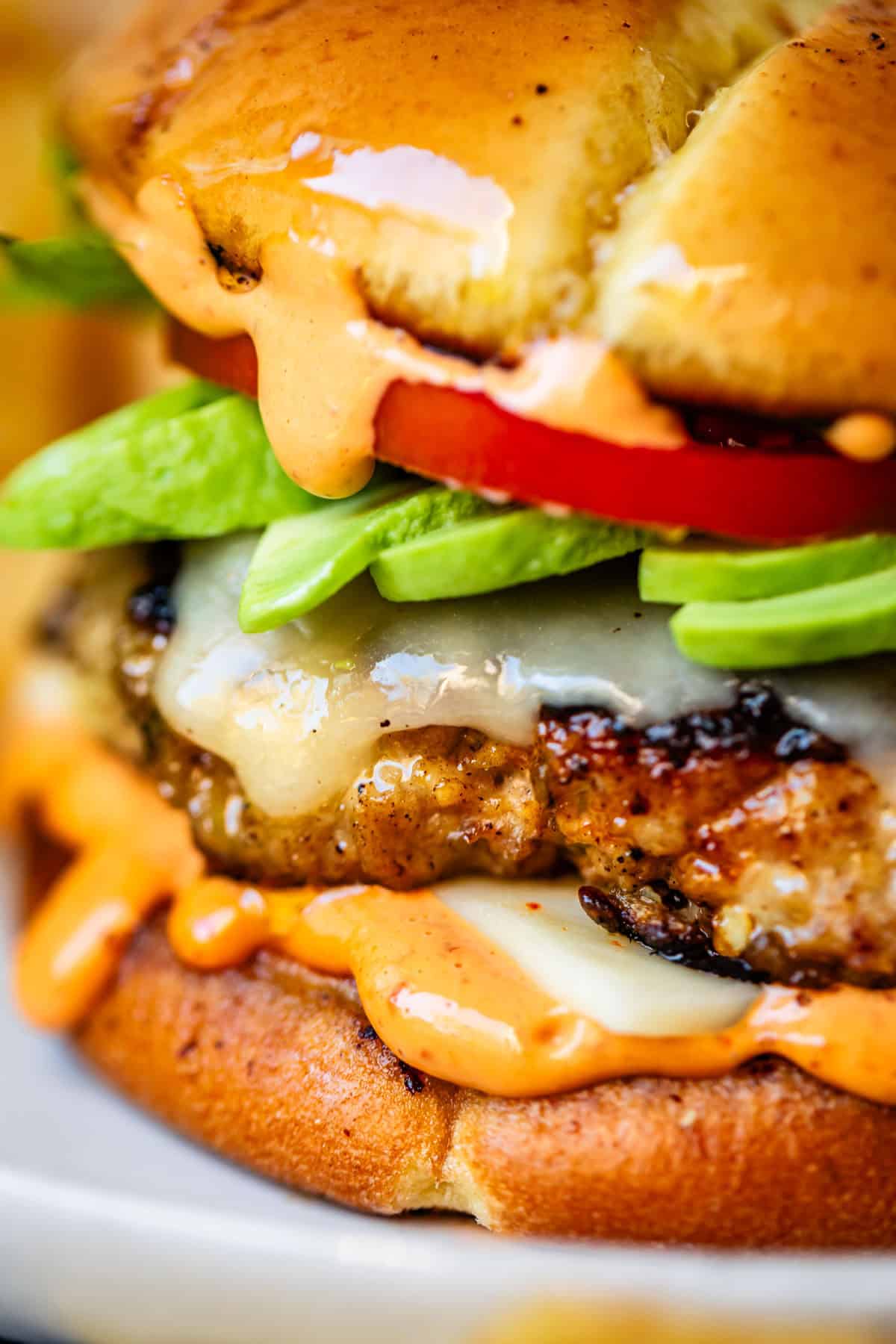 Close-up of a juicy cheeseburger with melted cheese, slices of avocado, tomato, and creamy orange sauce on a toasted bun. The burger is stacked high, highlighting fresh ingredients and dripping sauces.