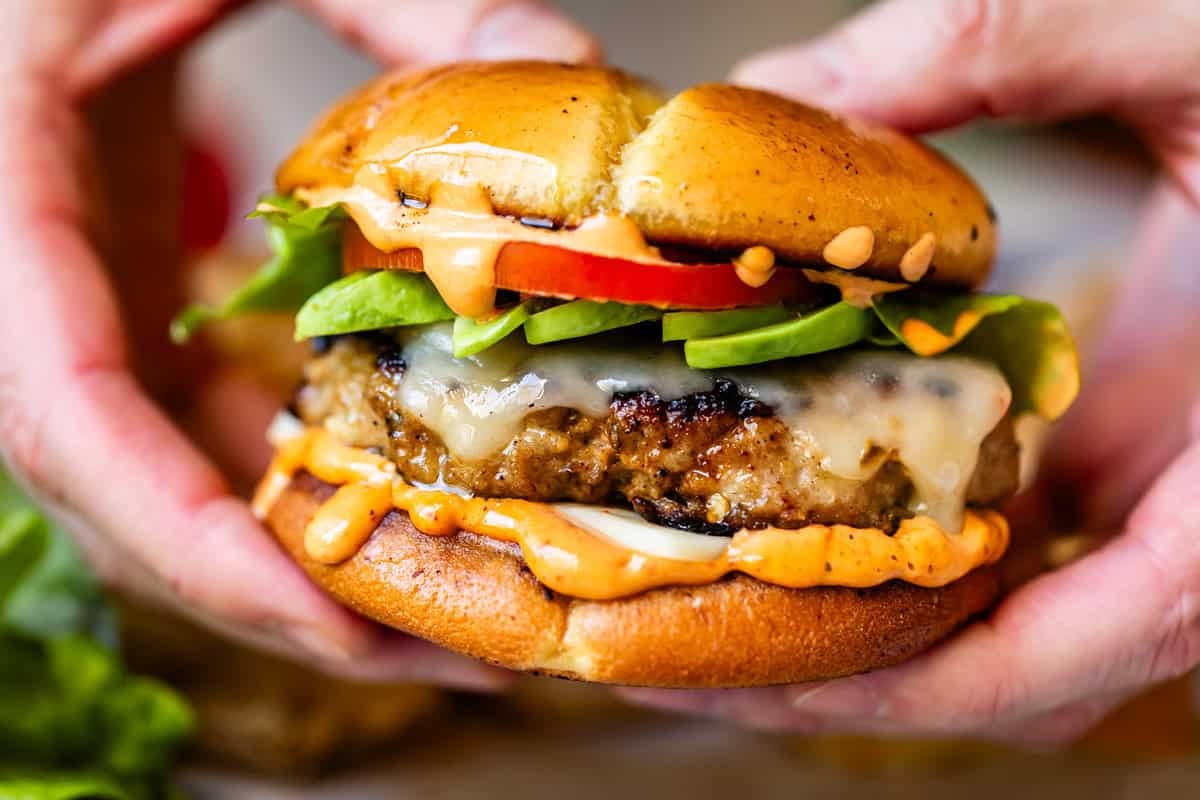 Close-up of hands holding a juicy cheeseburger with melted cheese, sliced avocado, lettuce, tomato, and creamy sauce on a toasted bun.
