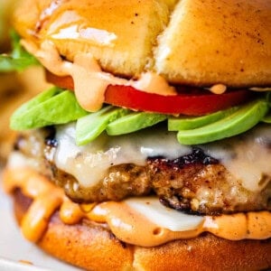 A close-up of a juicy cheeseburger with melted cheese, sliced avocado, tomato, lettuce, and a creamy orange sauce, all inside a glossy toasted bun.