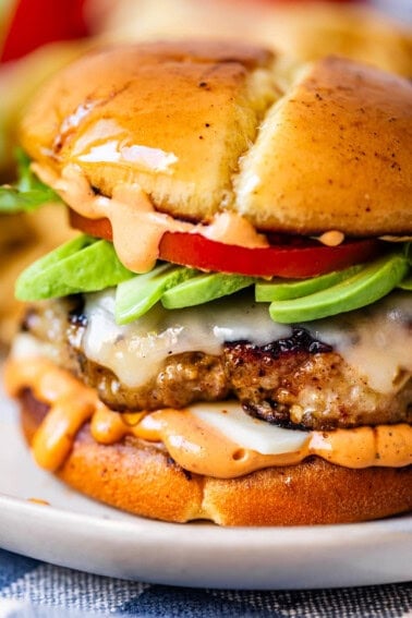 A close-up of a juicy cheeseburger with melted cheese, sliced avocado, tomato, lettuce, and a creamy orange sauce, all inside a glossy toasted bun.