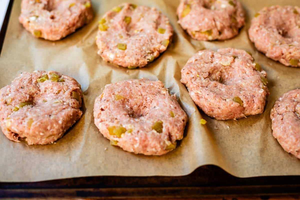 Raw, seasoned ground meat patties with chopped vegetables are arranged on a baking sheet lined with parchment paper, ready to be cooked. Each patty has a small indentation in the center.
