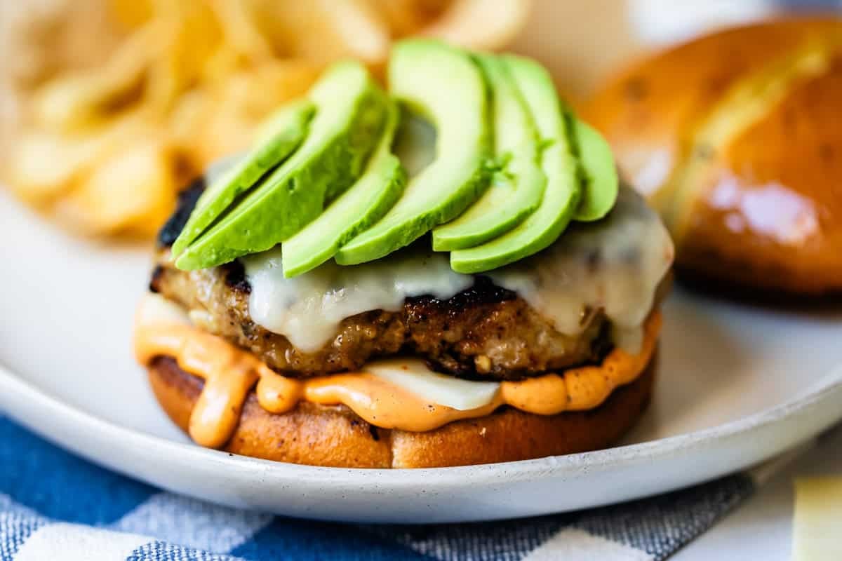 A close-up of a grilled burger patty topped with melted cheese and sliced avocado, on a toasted bun with sauce, served on a plate with potato chips in the background.