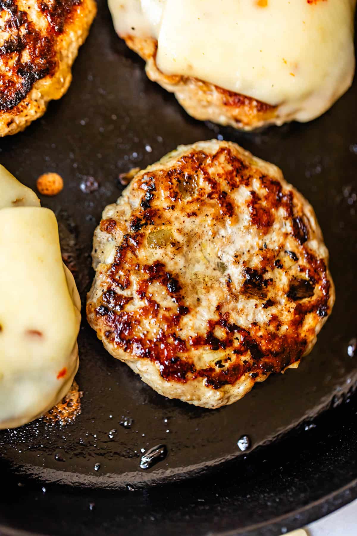 A close-up of turkey burger patties cooking in a skillet, with some patties topped with melted cheese and the surface showing crispy, browned grill marks.