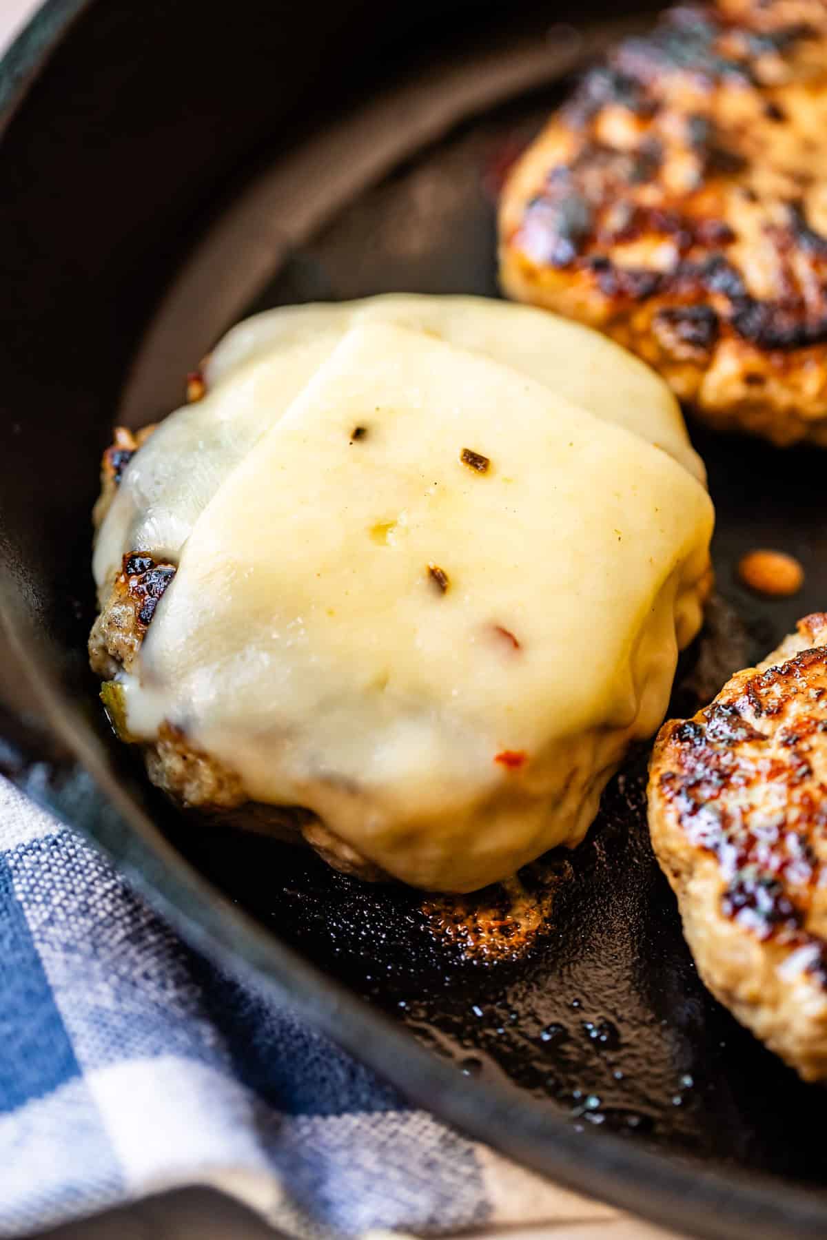 Close-up of a cooked burger patty topped with melted cheese in a black skillet, next to another plain patty, with part of a blue and white cloth visible in the corner.