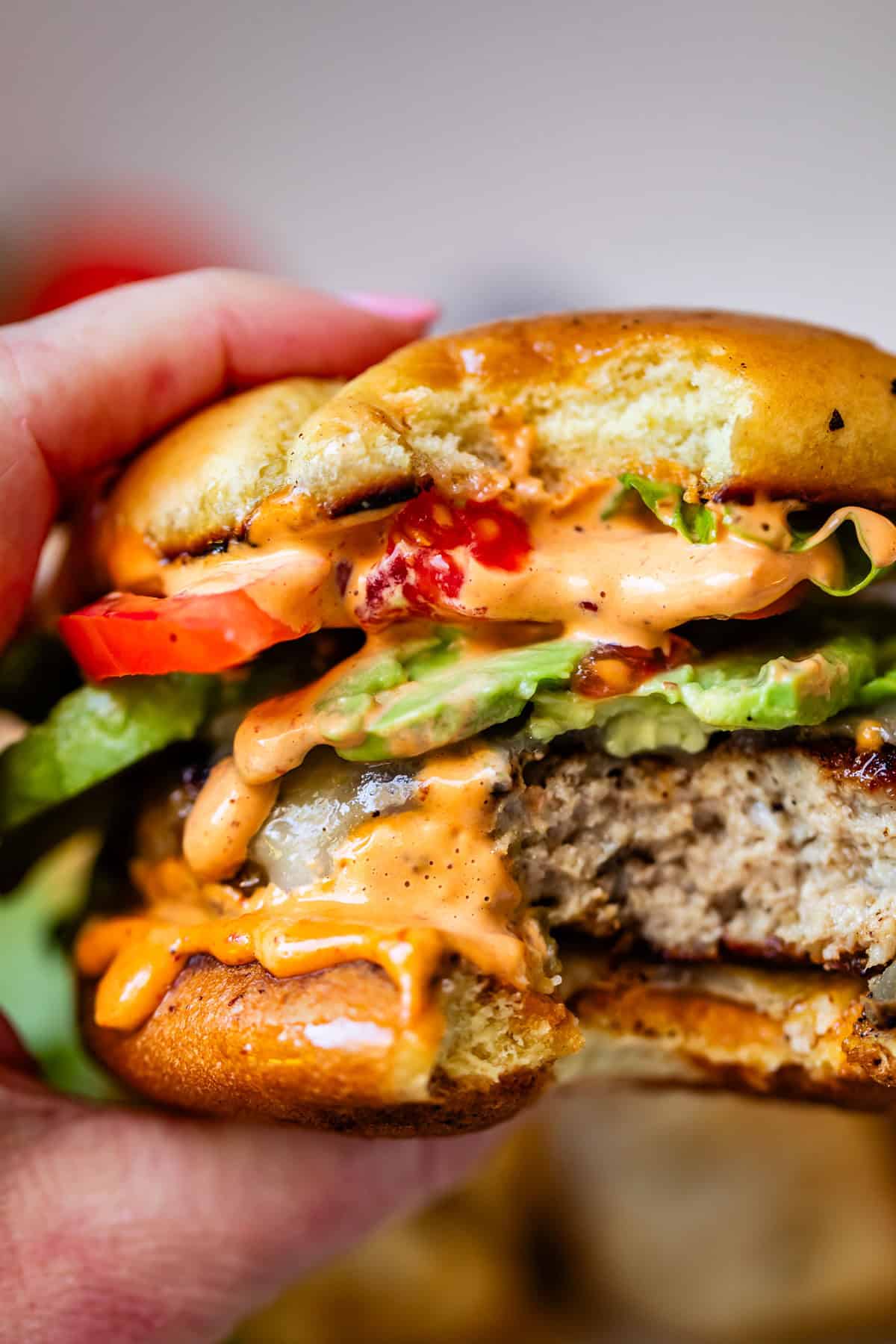 A hand holds a bitten burger with lettuce, tomato, melted cheese, and creamy sauce dripping out, revealing a juicy patty and a toasted bun.