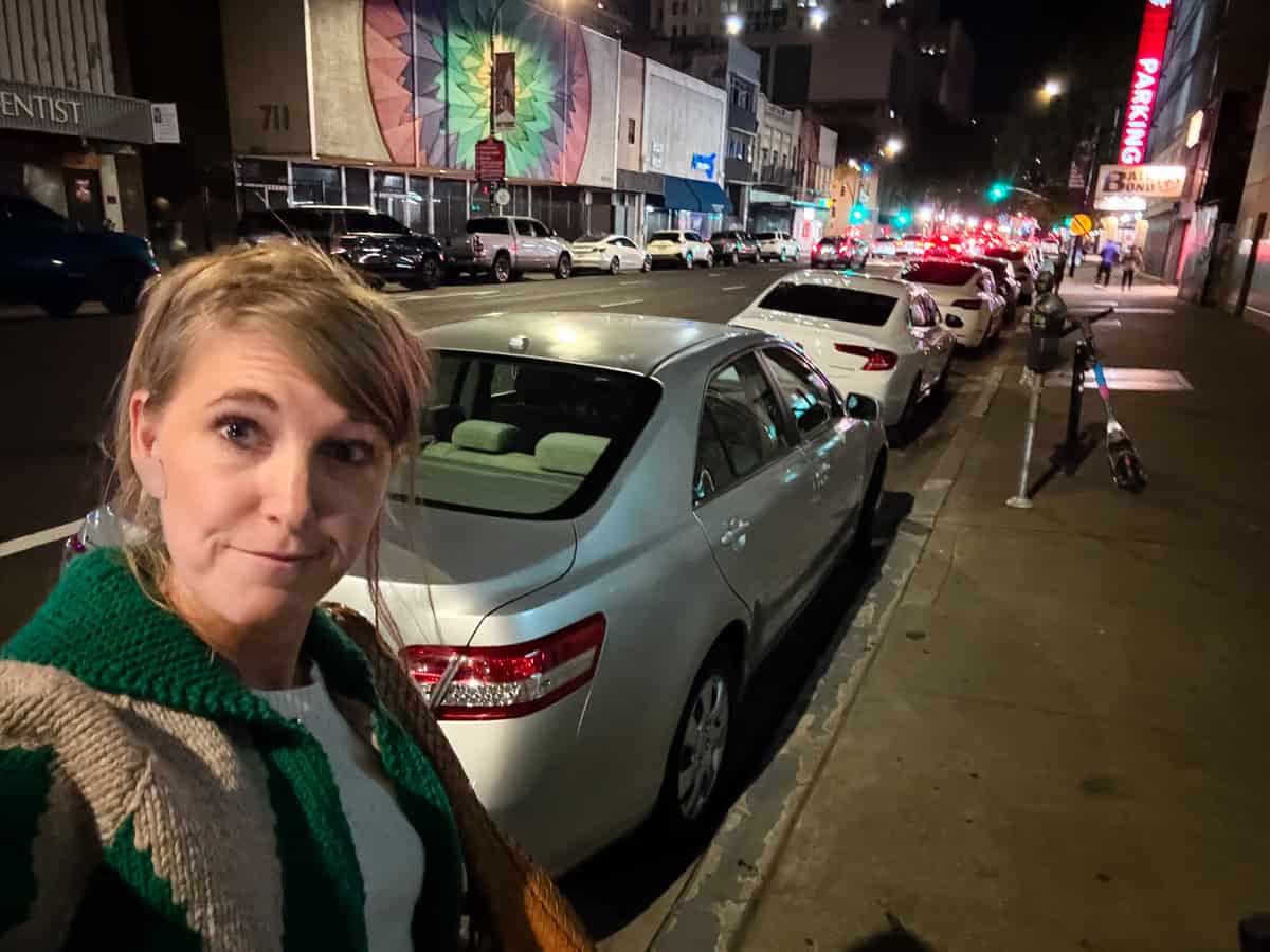 A woman takes a selfie on a city sidewalk at night, with parked cars lining the street, a colorful mural behind her, and bright streetlights illuminating the scene.
