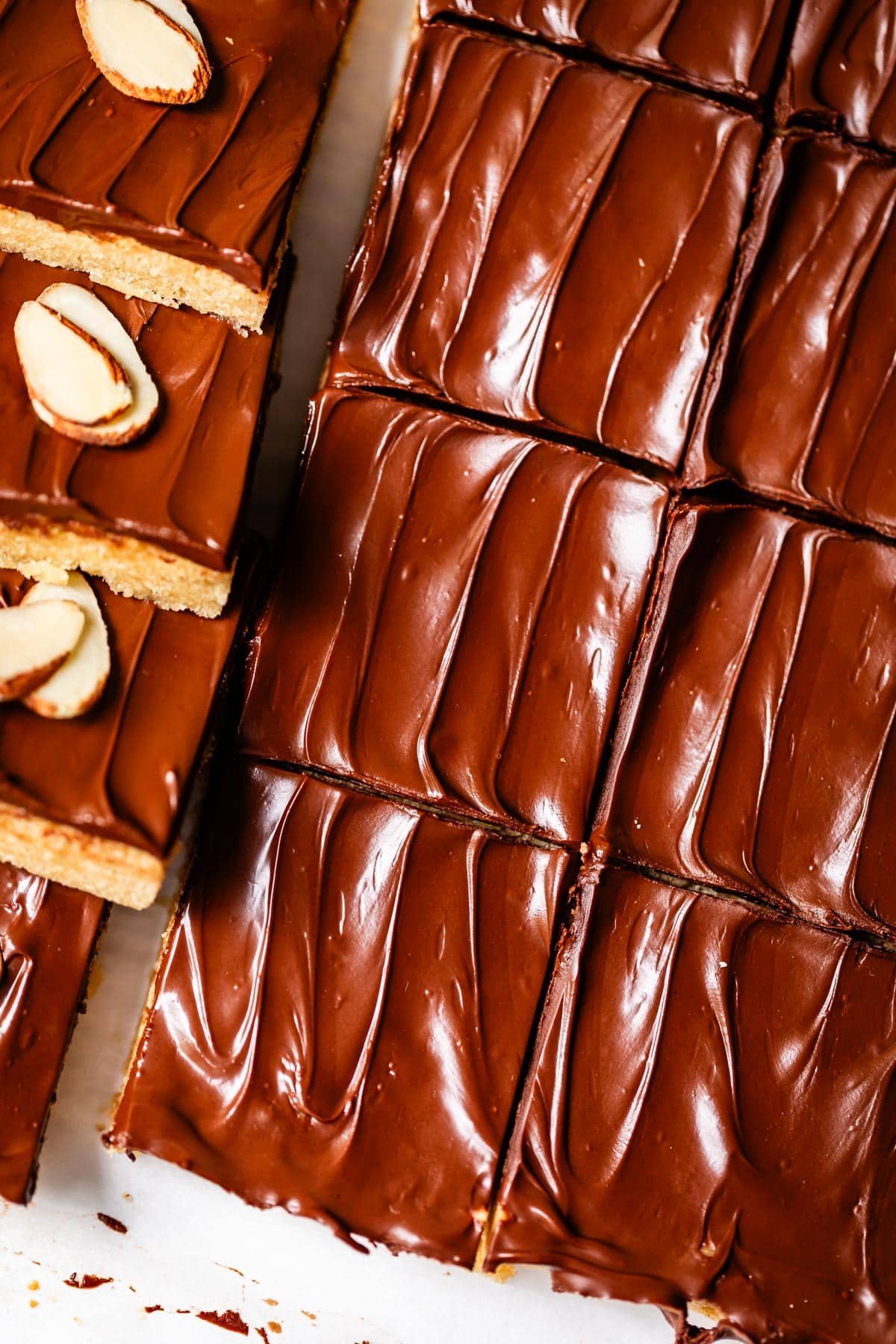 Close-up of chocolate-covered dessert bars, sliced into squares. Some bars are topped with almond slices, while most have a smooth, glossy chocolate layer with visible swirls.
