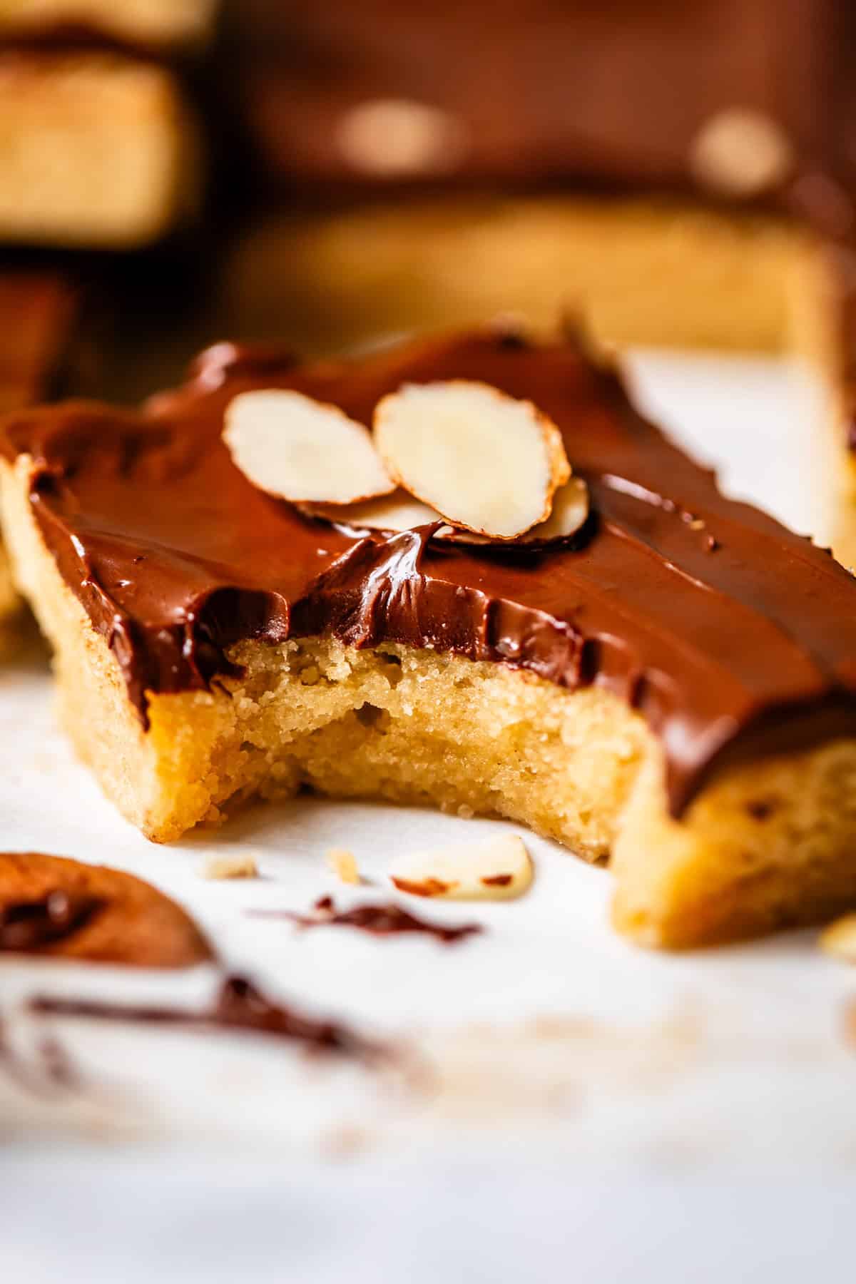 A close-up of a chocolate almond bar with a bite taken out, topped with sliced almonds. The bar has a golden, soft interior and a thick layer of glossy chocolate on top. Crumbs and chocolate pieces are scattered around.