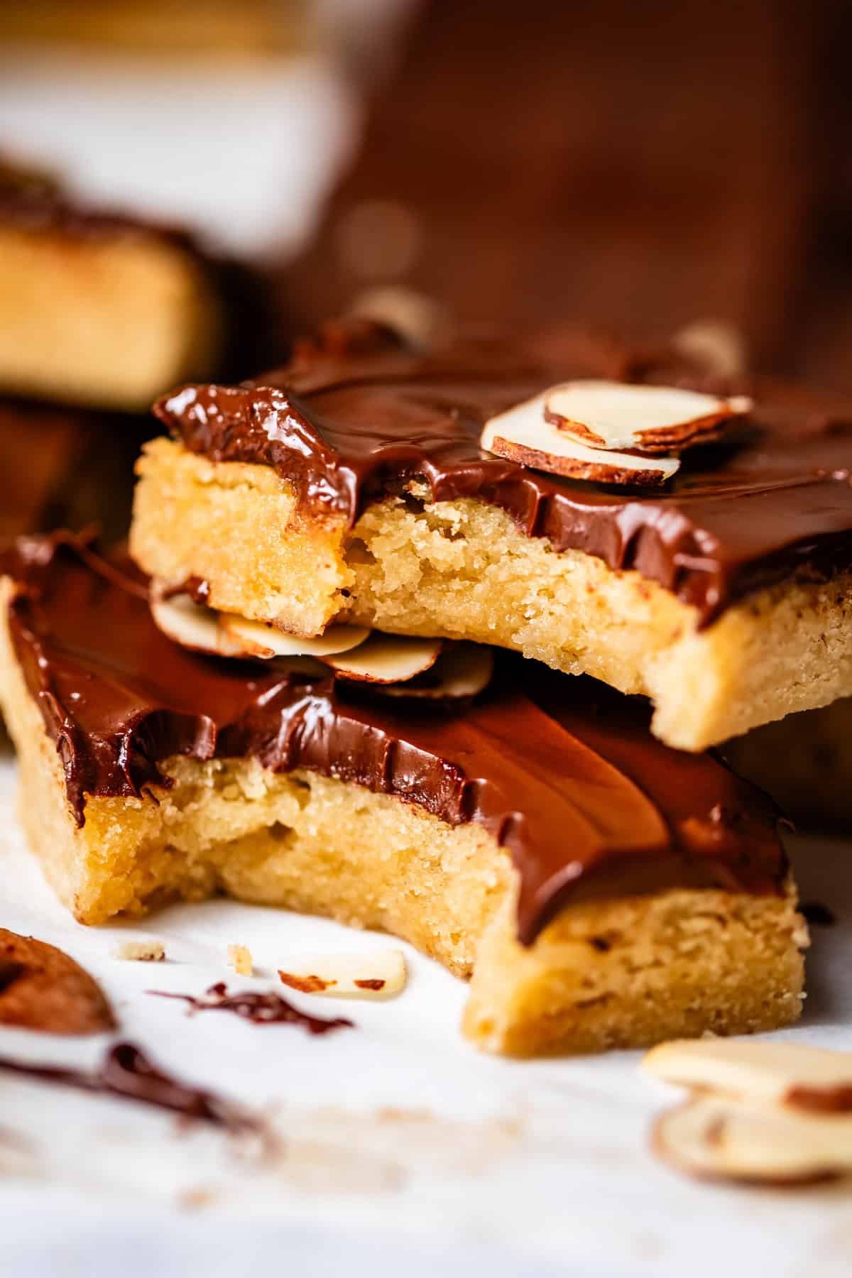 Close-up of two golden cookie bars with a thick layer of chocolate on top, garnished with sliced almonds. One bar has a bite taken out, revealing a soft, chewy interior.