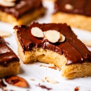 A close-up of a chocolate-topped bar cookie garnished with sliced almonds. One bar in the foreground has a bite taken out, revealing a soft, golden interior. Other bars and scattered almond slices are in the background.