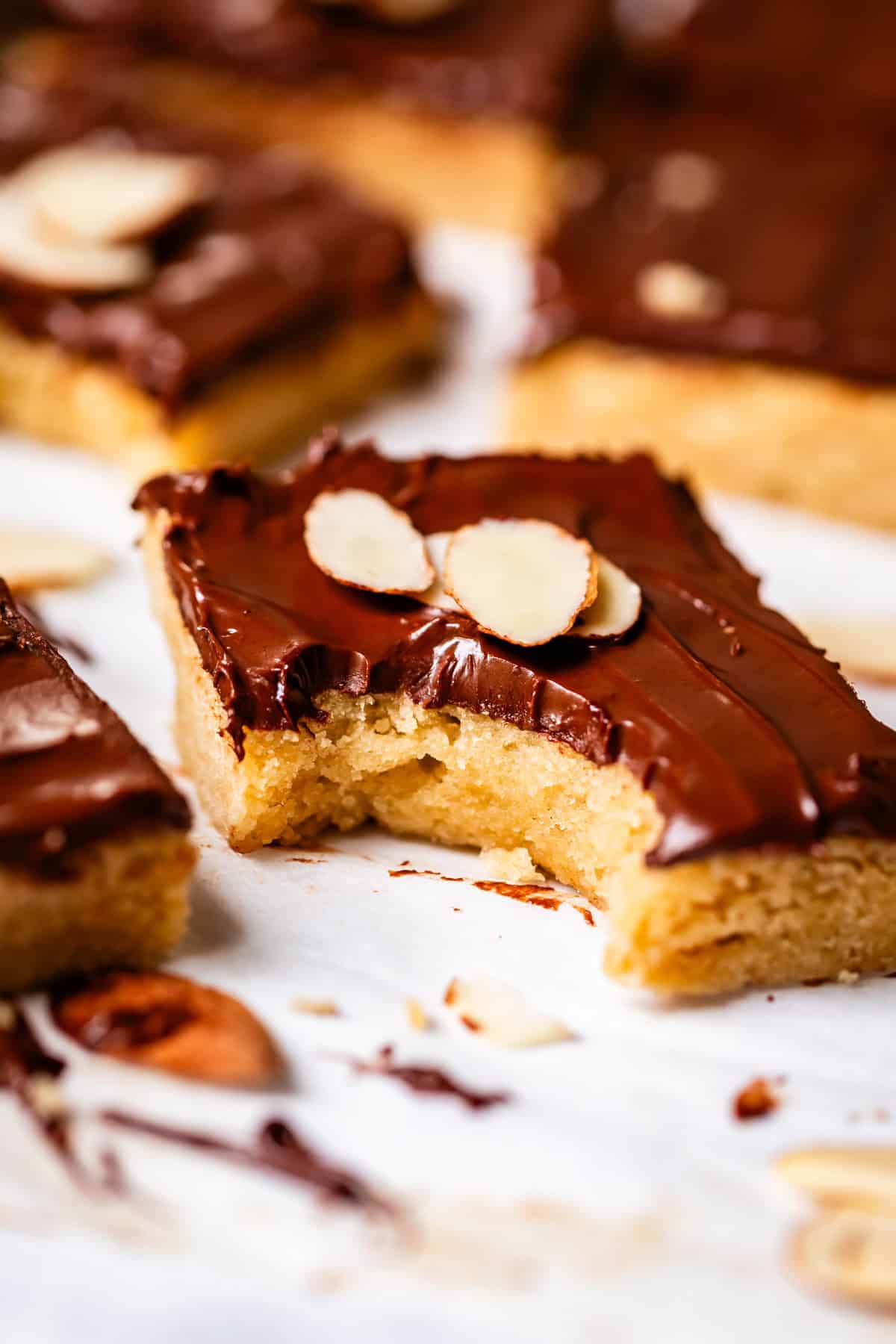 A close-up of a chocolate-topped bar cookie garnished with sliced almonds. One bar in the foreground has a bite taken out, revealing a soft, golden interior. Other bars and scattered almond slices are in the background.