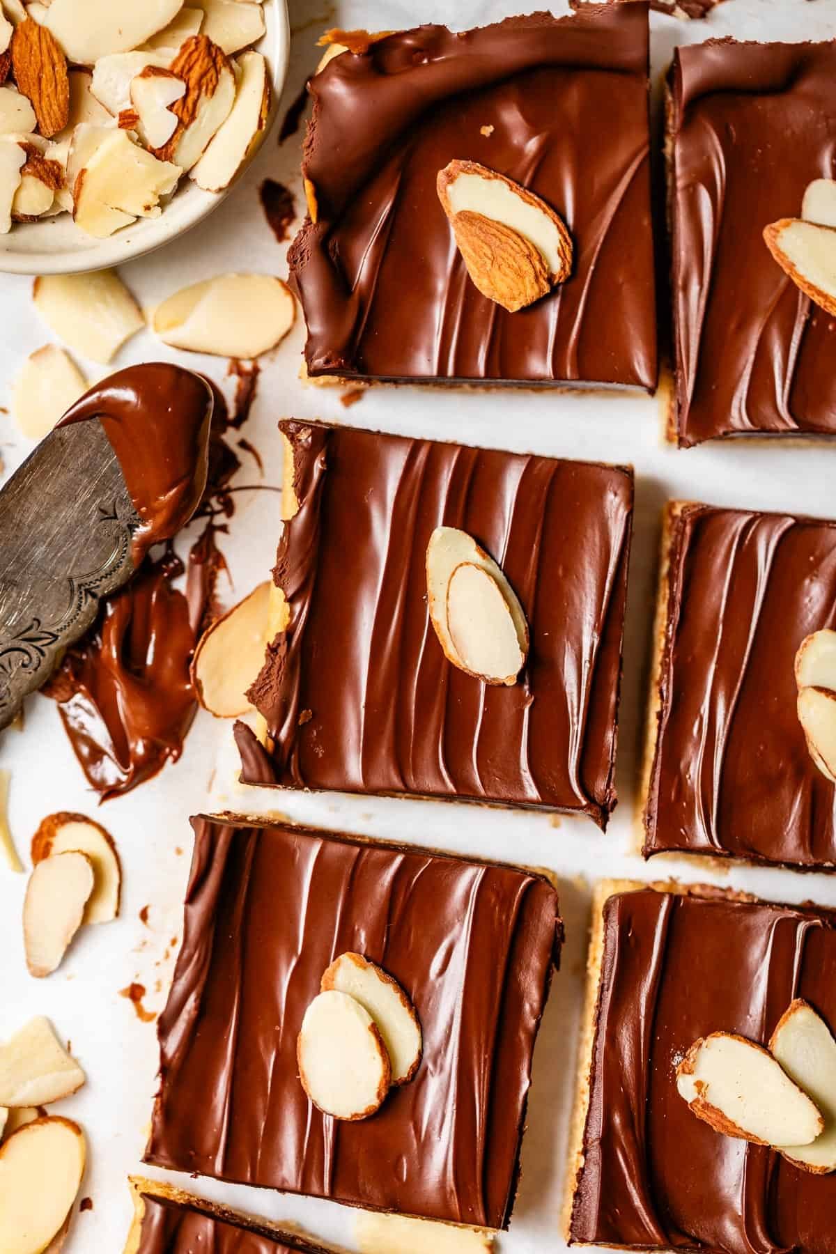 Six chocolate-covered bars topped with sliced almonds are arranged on a white surface. A knife with chocolate and scattered almond slices appear on the left, with a small bowl of almonds in the top corner.