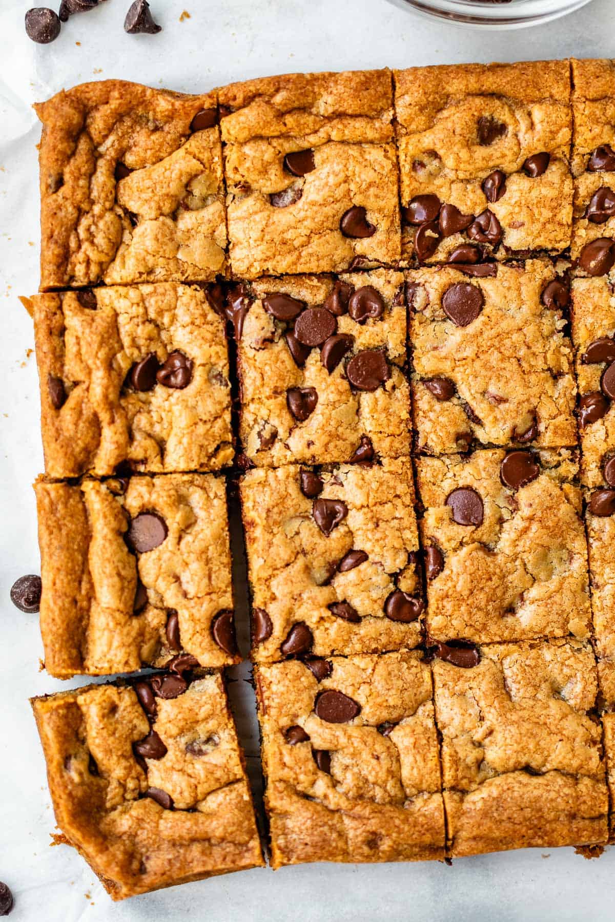 A batch of chocolate chip cookie bars cut into squares on a white surface, with a few chocolate chips scattered nearby.