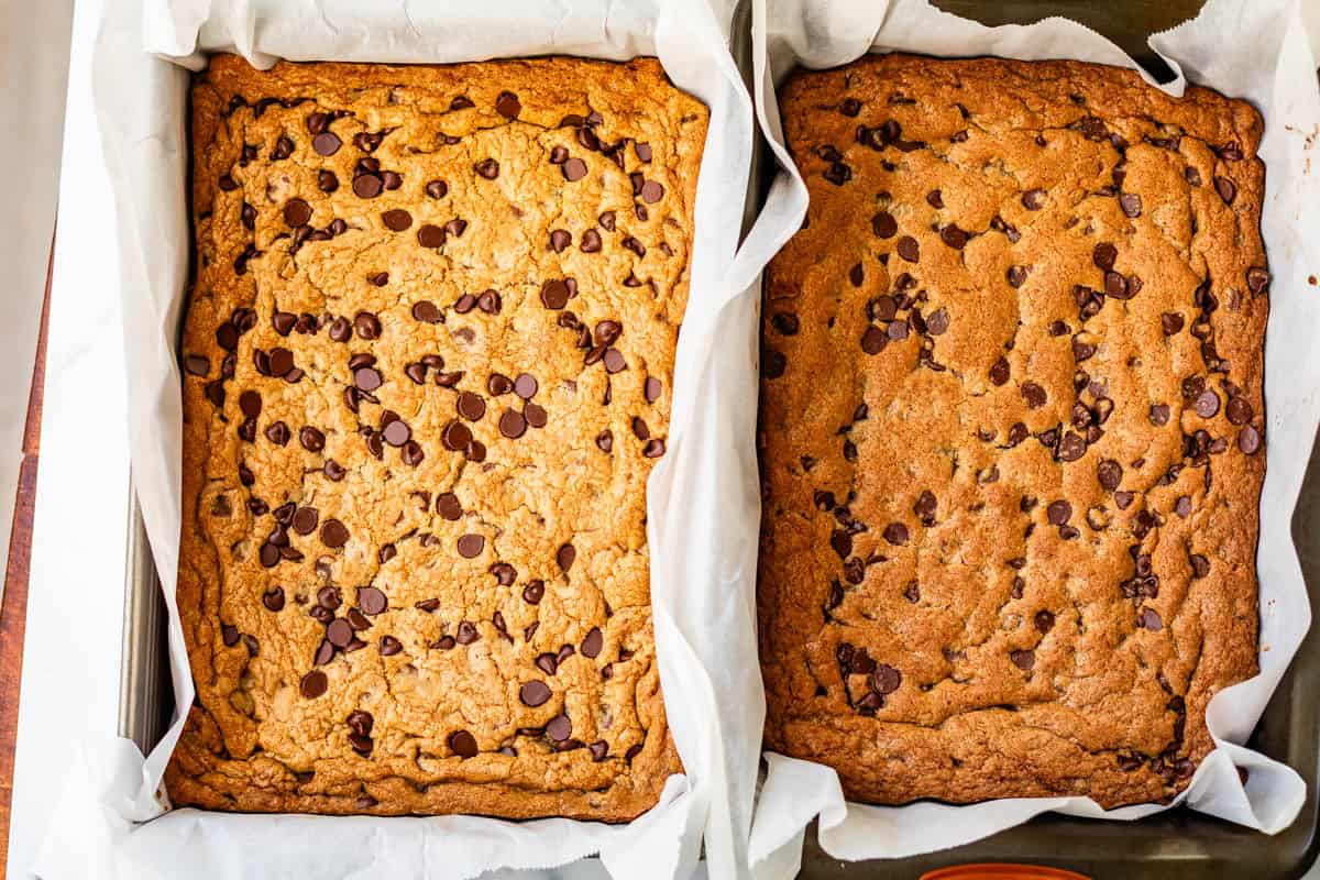 Two rectangular pans lined with parchment paper, each filled with golden-brown chocolate chip cookie bars, cooling and ready to be cut. Chocolate chips are scattered on top of the bars.