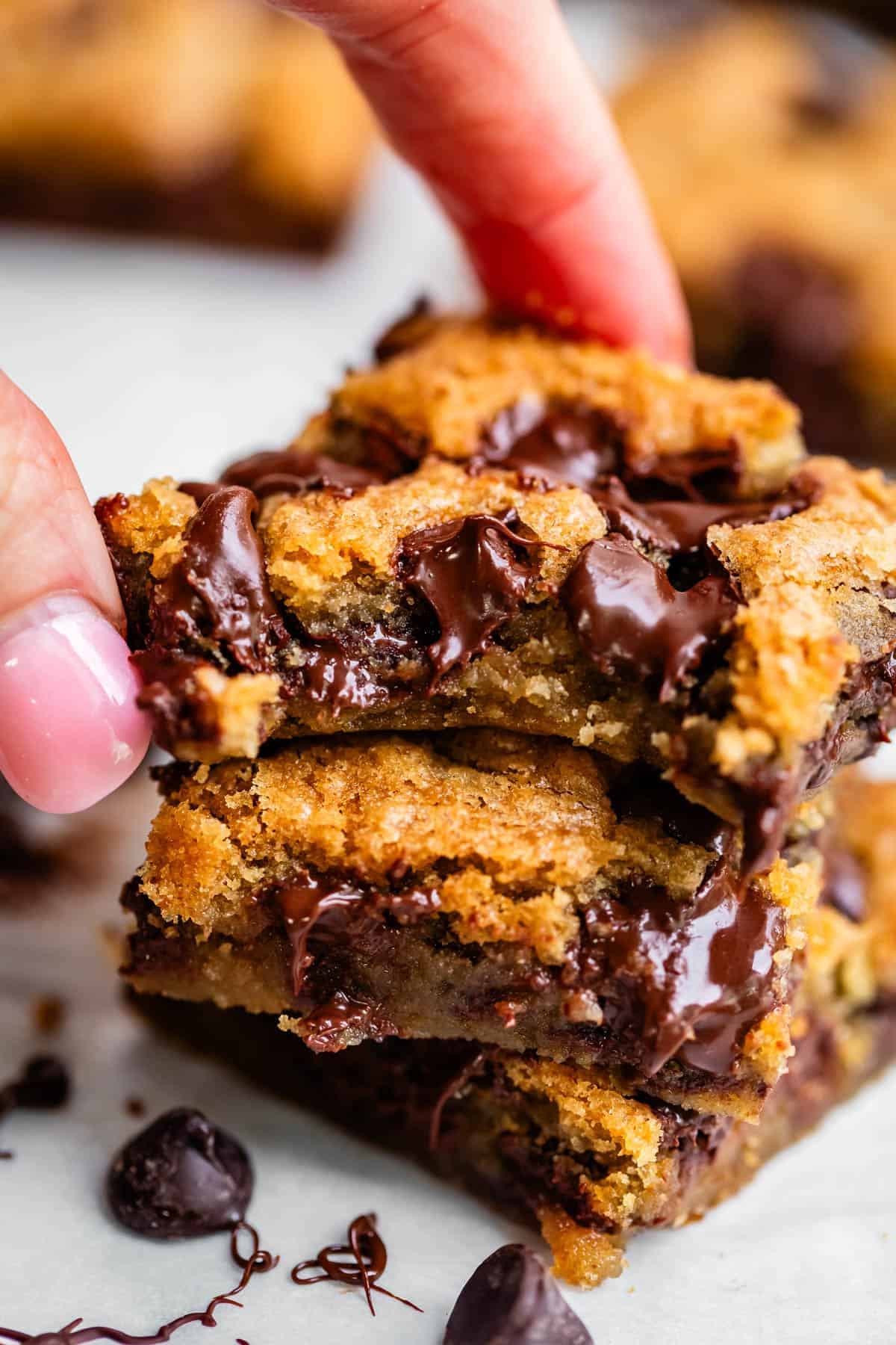 A close-up of a hand holding a stack of gooey chocolate chip cookie bars, with melted chocolate chips oozing from the soft, golden-brown centers.