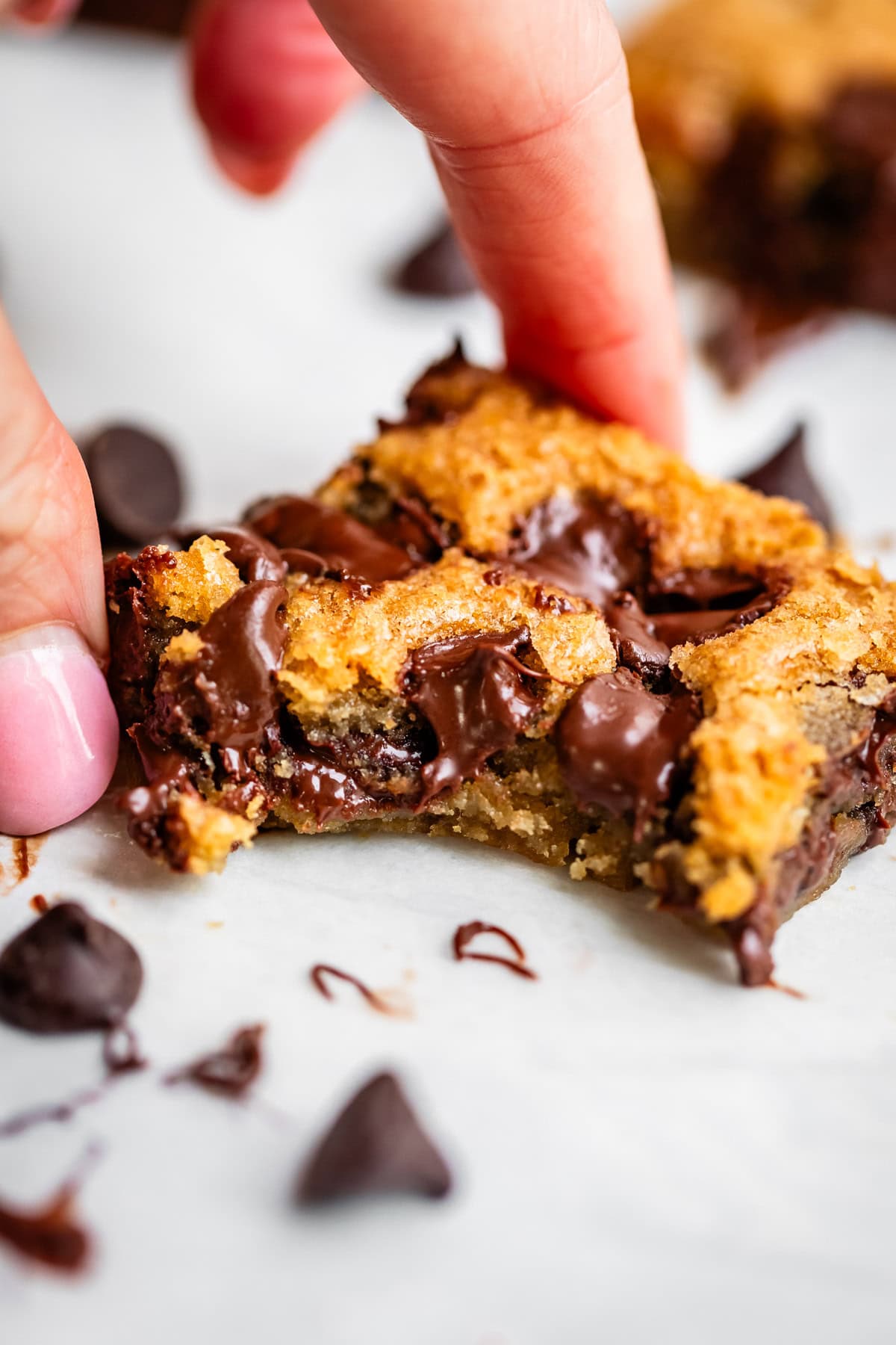 A hand holding a gooey, golden-brown chocolate chip cookie bar with melted chocolate chips, surrounded by chocolate chips on a white surface.