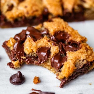 A close-up of a gooey chocolate chip cookie bar with melted chocolate chips, featuring a bite taken out. More bars and chocolate chips are visible in the background on a white surface.