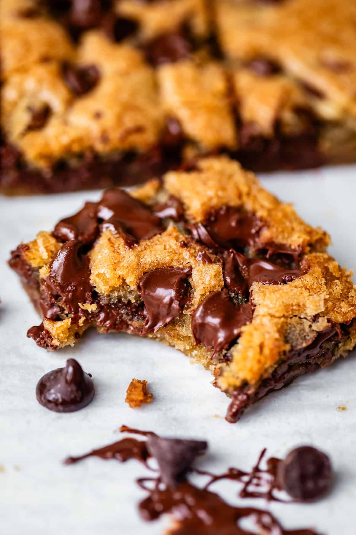 A close-up of a gooey chocolate chip cookie bar with melted chocolate chips, featuring a bite taken out. More bars and chocolate chips are visible in the background on a white surface.