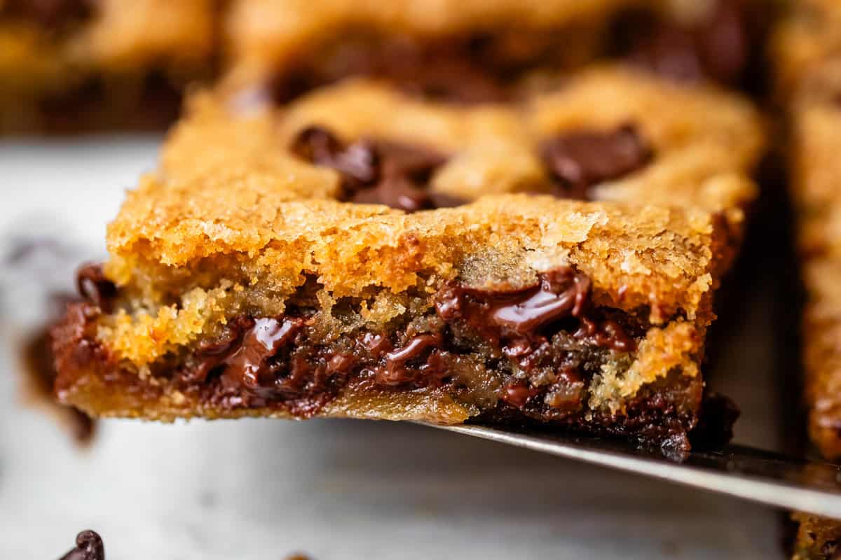 A close-up of a gooey chocolate chip cookie bar with melted chocolate chunks, golden brown edges, and a soft, chewy interior on a spatula.
