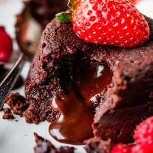 A close-up of a chocolate lava cake with melted chocolate oozing out, topped with a fresh strawberry. The dessert is served on a plate with a spoon and raspberries nearby.