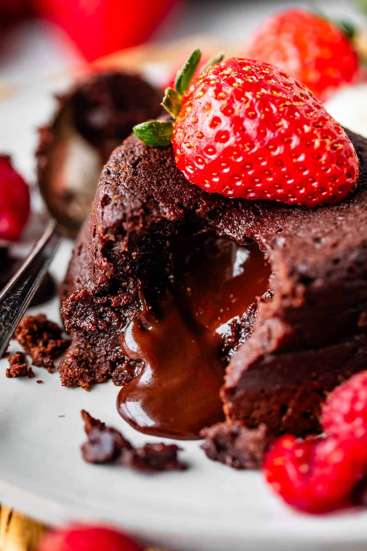 A close-up of a chocolate lava cake with melted chocolate oozing out, topped with a fresh strawberry. The dessert is served on a plate with a spoon and raspberries nearby.