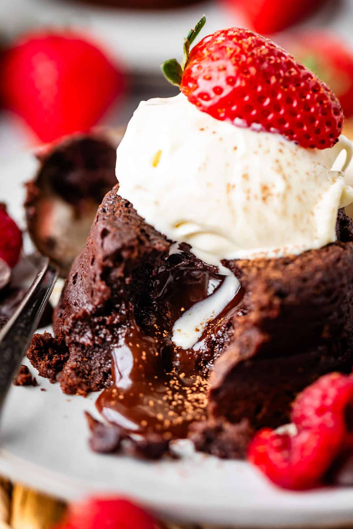 A close-up of a chocolate lava cake with melted chocolate oozing out, topped with a scoop of vanilla ice cream and a fresh strawberry, surrounded by more strawberries and raspberries on a plate.