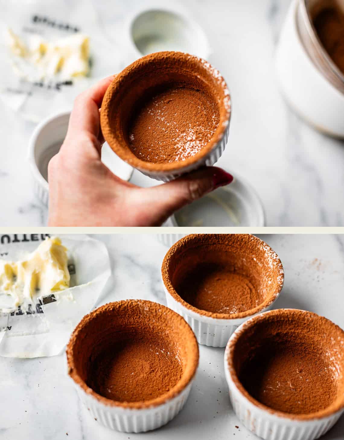A hand holds a small white ramekin coated with cocoa powder. Below, two more ramekins are similarly coated, sitting on a marble surface near a butter wrapper and a stick of butter.