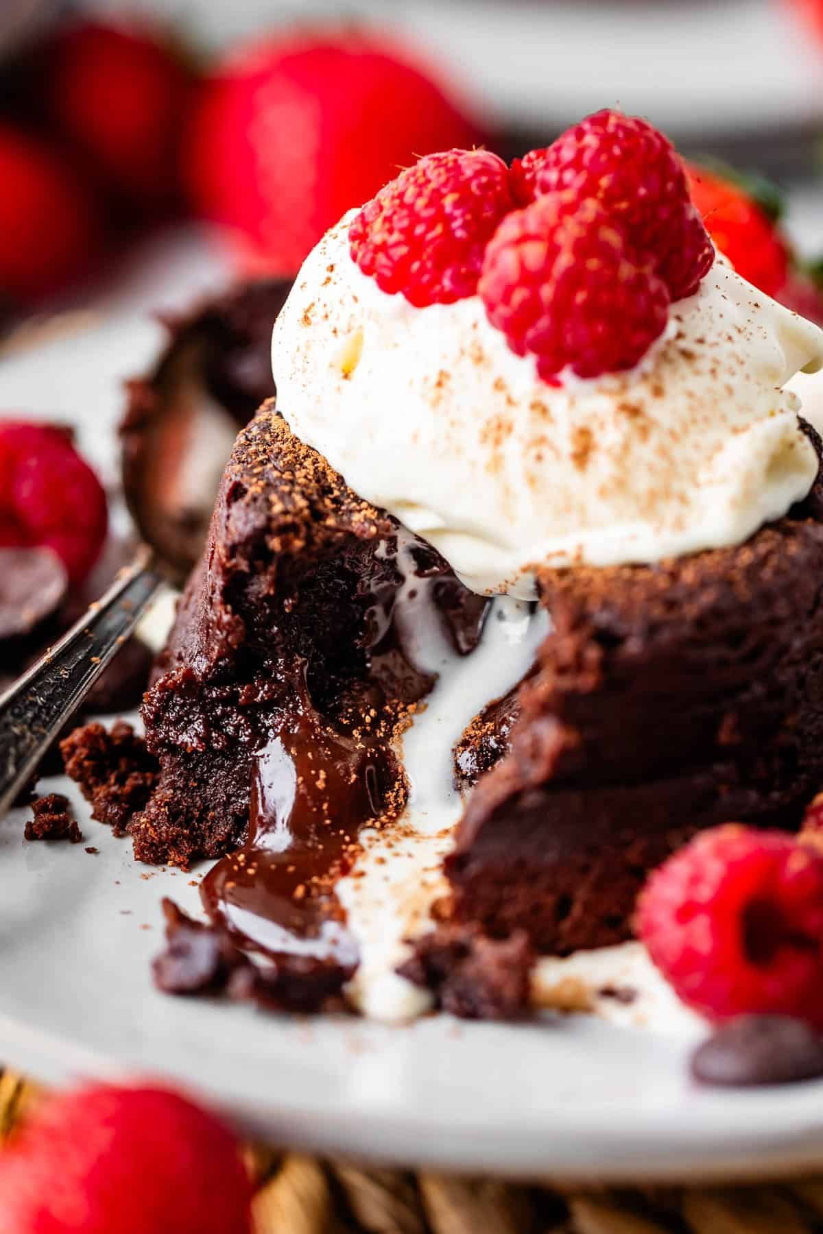 A rich chocolate lava cake with molten center, topped with whipped cream and fresh raspberries, sits on a white plate with a fork and scattered raspberries nearby.