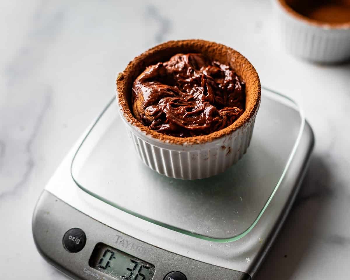 A small white ramekin filled with chocolate cake batter sits on a digital kitchen scale, displaying a weight of 3.5 ounces; another ramekin is blurred in the background on a white surface.
