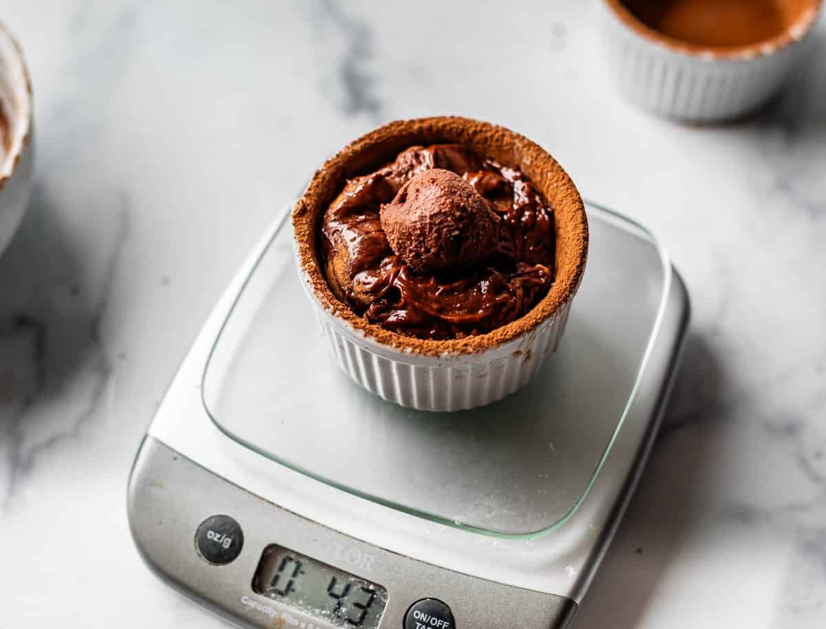 A chocolate souffl&eacute; in a white ramekin sits on a digital kitchen scale displaying 4.43 ounces, on a marble countertop. Another ramekin and a mixing bowl are partially visible in the background.