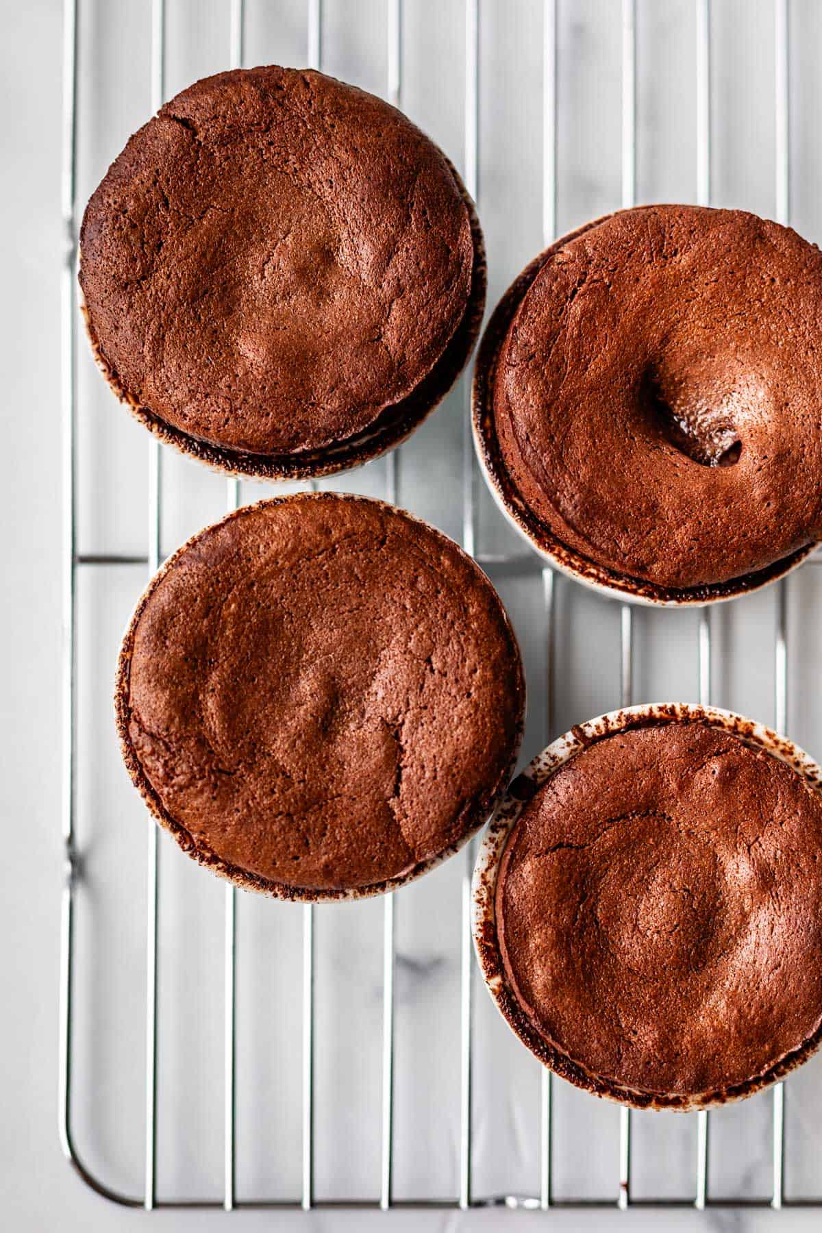 Four baked chocolate souffl&eacute;s in white ramekins cooling on a metal wire rack, seen from above. The souffl&eacute;s have risen tops with a slightly cracked, rich brown surface.