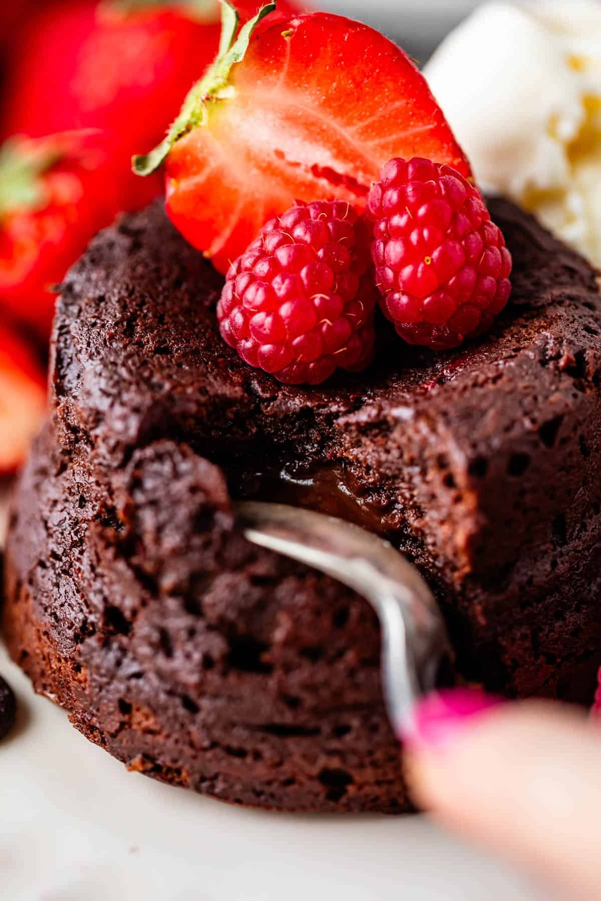 Close-up of a chocolate lava cake with a molten center, topped with fresh raspberries and a halved strawberry. A fork is breaking into the cake. In the background, there are more strawberries and a scoop of vanilla ice cream.