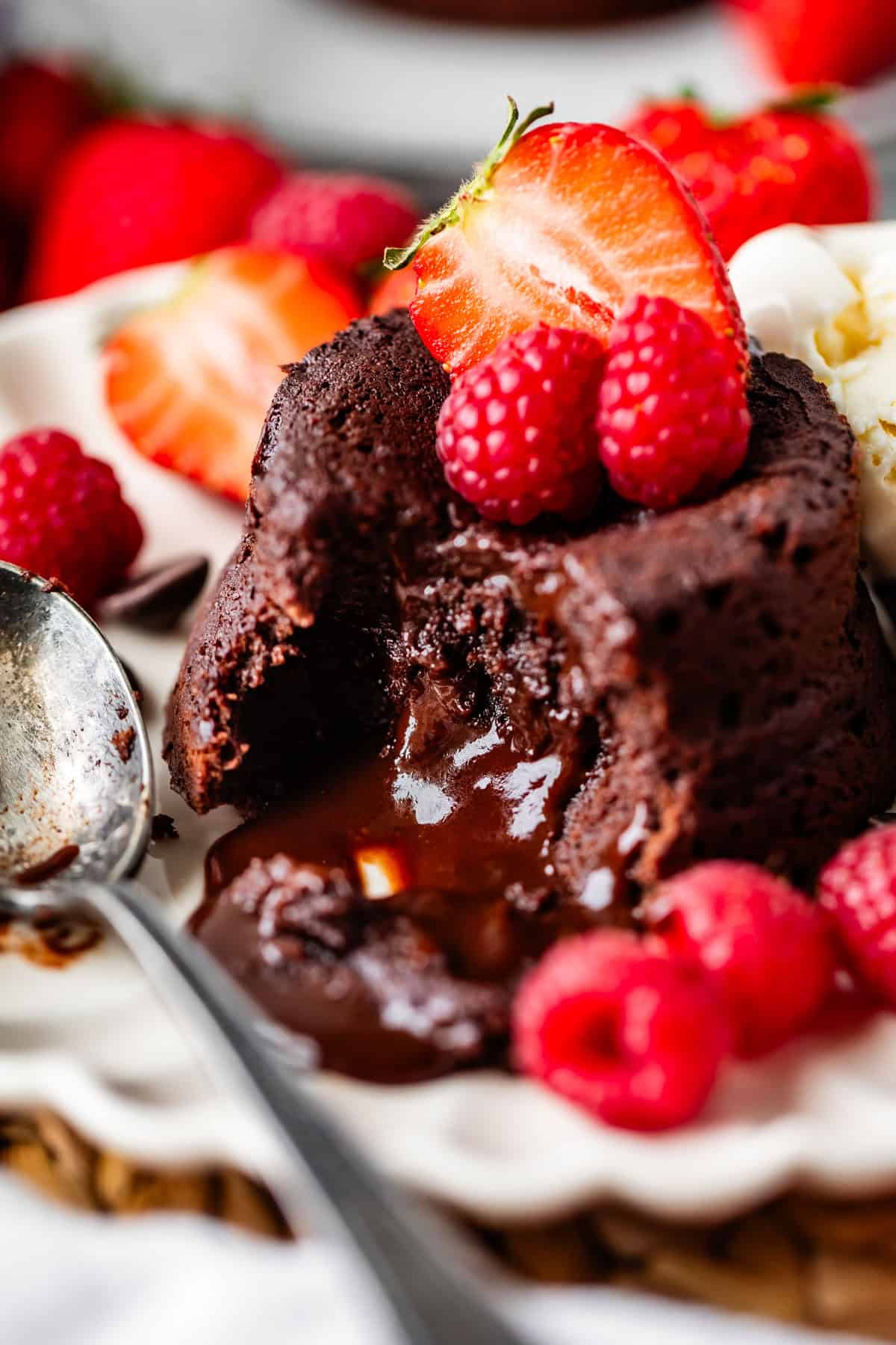 A rich chocolate lava cake with a gooey molten center is topped with fresh raspberries and a halved strawberry, served on a white plate with a spoon and more berries on the side.