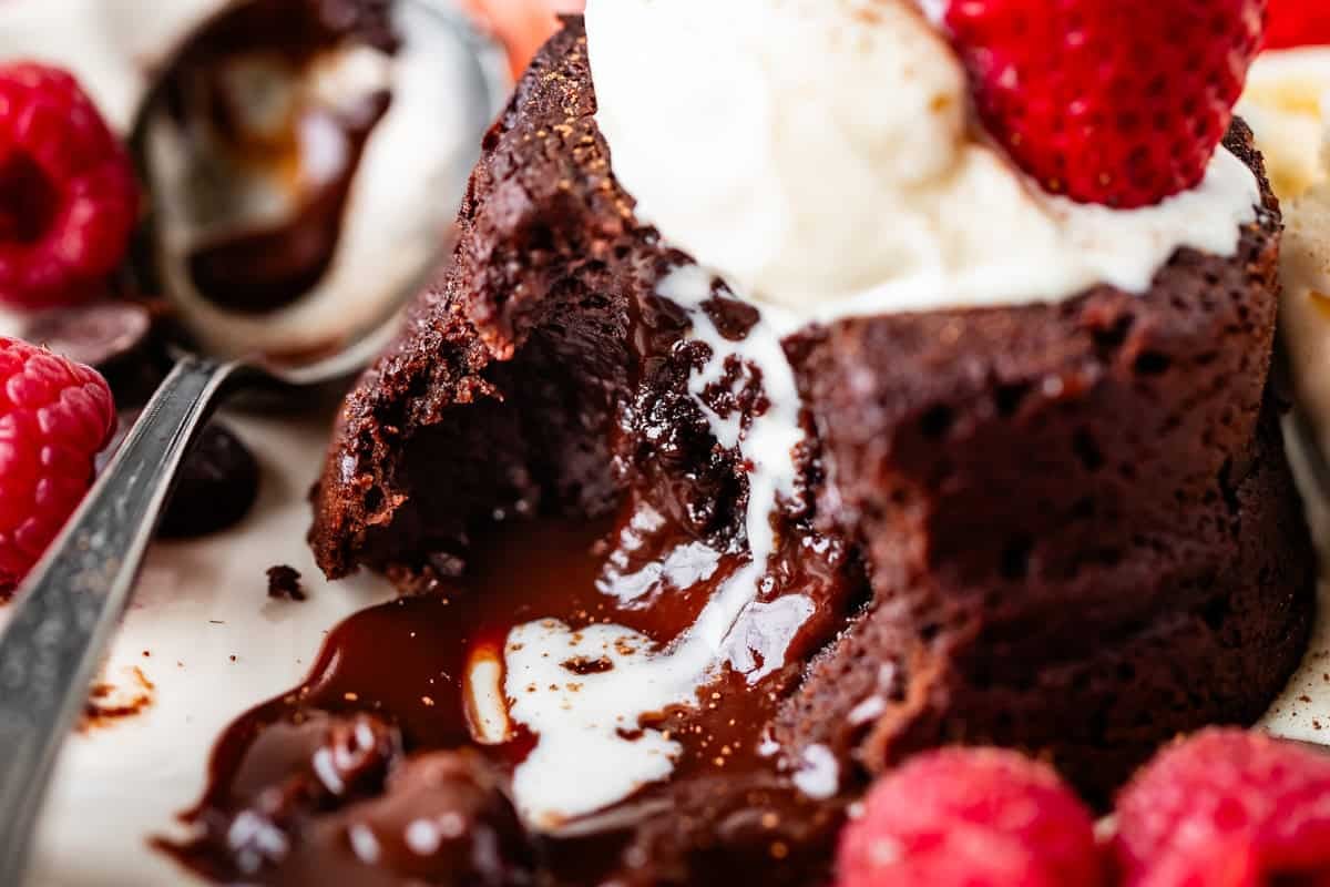 A close-up of a molten chocolate lava cake with a gooey chocolate center, topped with vanilla ice cream and a strawberry, surrounded by fresh raspberries and a spoon scooping the dessert.