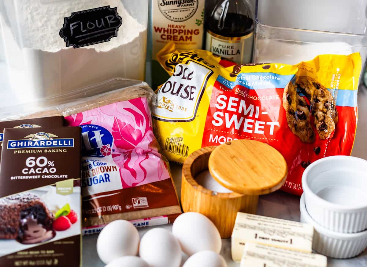 A variety of baking ingredients including flour, heavy cream, vanilla, brown sugar, chocolate chips, butter, eggs, and a bar of 60% cacao bittersweet chocolate arranged on a counter.